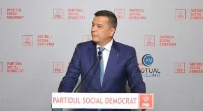 Un bărbat într-un costum albastru și cravată deschisă stă la un pupitru în fața unui fundal cu logourile PSD repetate.