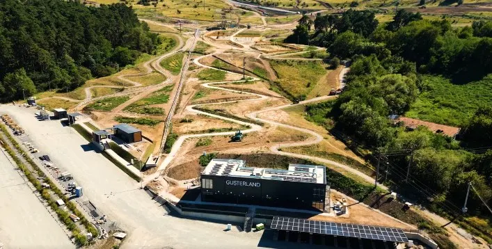O vedere aeriană prezintă un teren de schi în construcție cu piste de pământ, un lift de tip telecabina și o clădire neagră modernă.