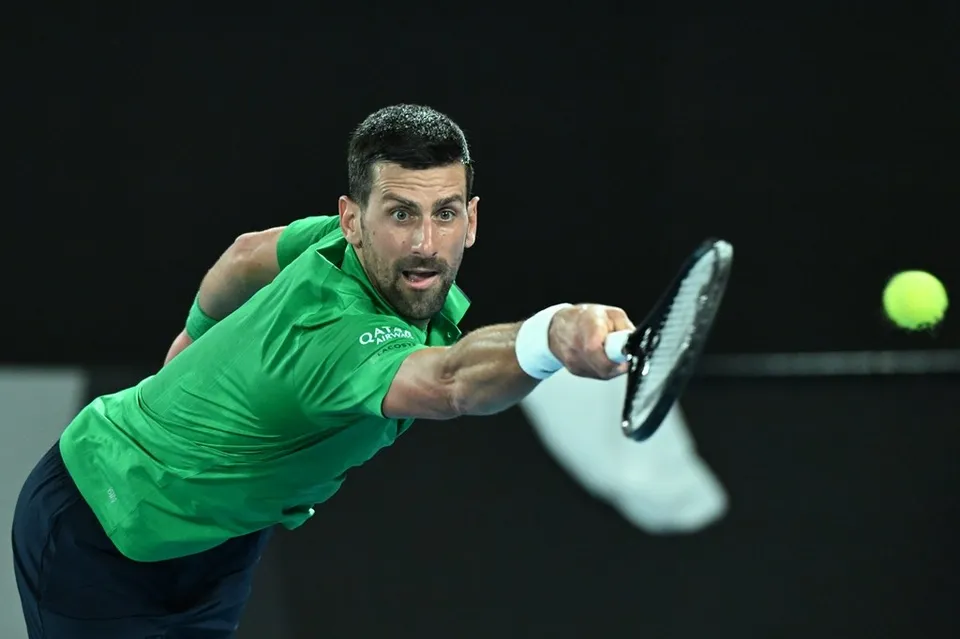 Un bărbat cu tricou verde și pantaloni închiși lovește o minge de tenis cu o rachetă într-o poziție de întindere.