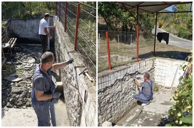 Un bărbat în cămașă gri și pantaloni de lucru lucrează la un zid de piatră lângă o structură de beton.
