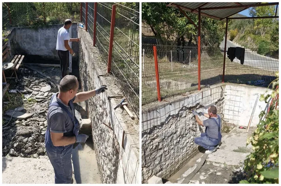 Un bărbat în cămașă gri și pantaloni de lucru lucrează la un zid de piatră lângă o structură de beton.