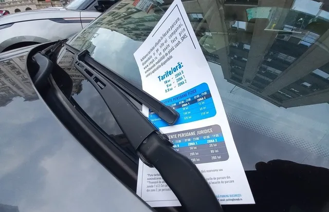 O foaie de hârtie cu text și tabele colorate este lipită pe parbrizul unei mașini, lângă o ștergătoare de parbriz neagră.