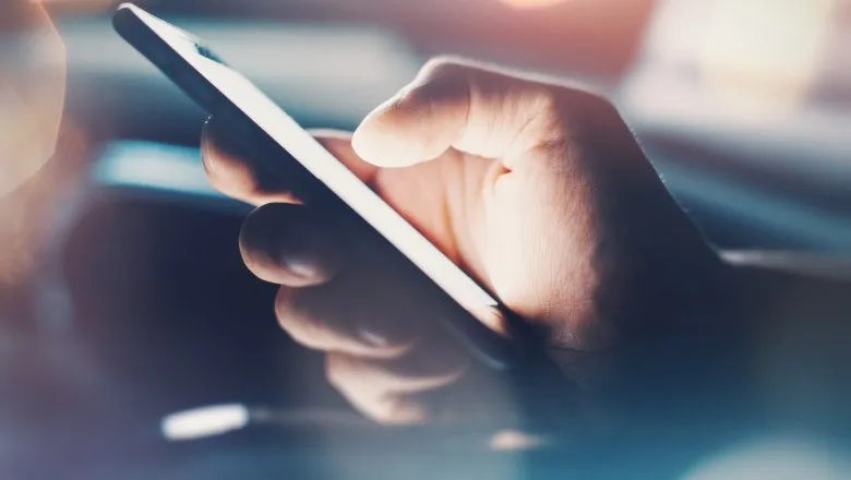 O persoană ține un smartphone negru cu ambele mâini într-un mediu cu lumină difuză.