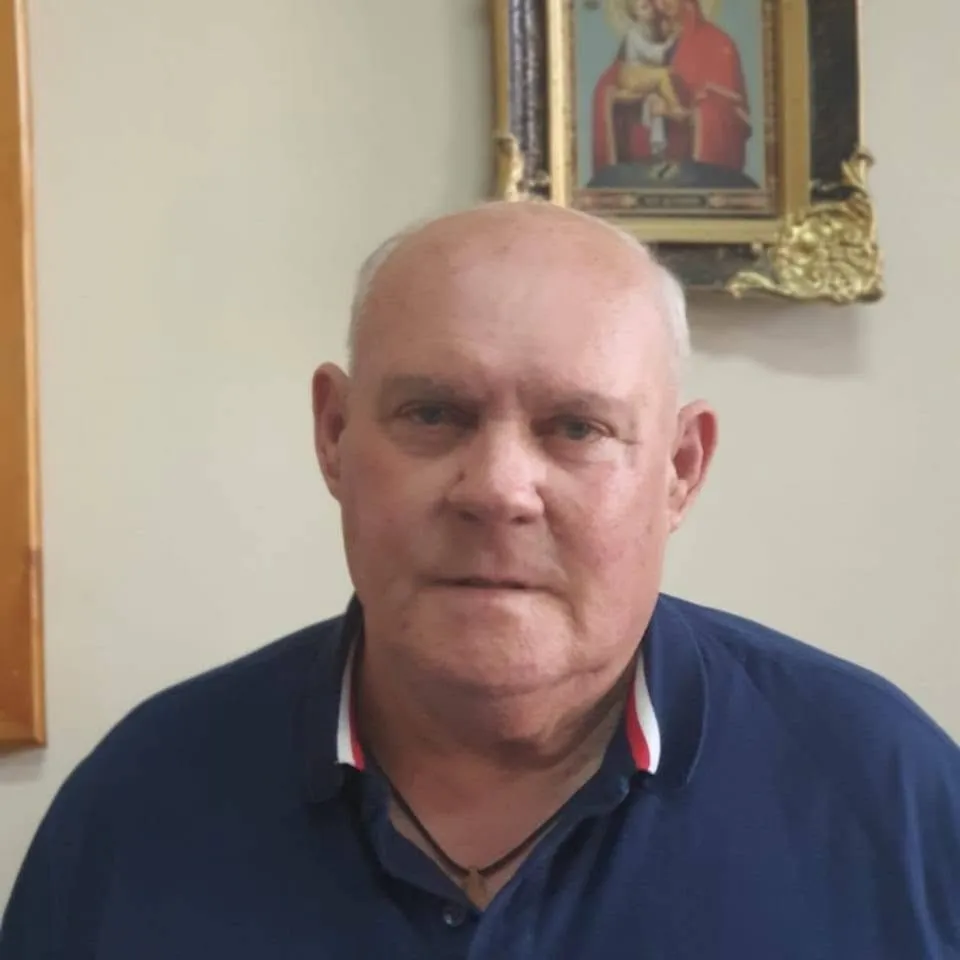 Un bărbat cu părul scurt și alb, purtând un tricou polo albastru, este poziționat în fața unui perete deschis cu un tablou în fundal.