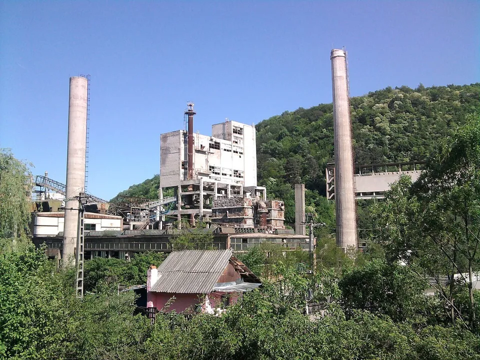 O fabrică cu două coșuri înalte de beton și clădiri industriale albe este situată într-o zonă cu vegetație abundentă și dealuri împădurite.