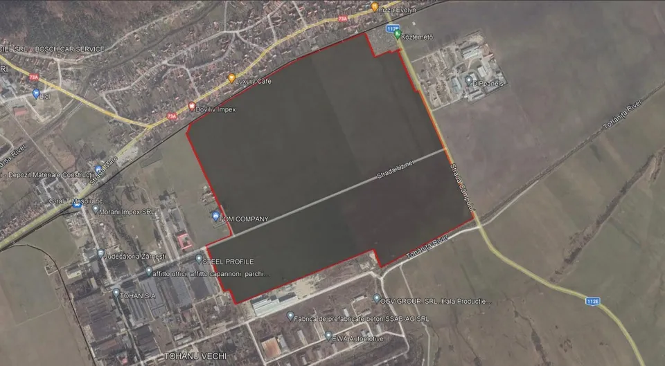 O vedere aeriană de satelit prezintă un teren mare, delimitat cu o linie roșie, situat lângă drumuri și clădiri industriale.