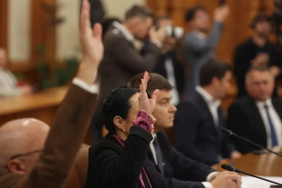 O femeie în costum negru și un bărbat alături ridici brațele în semn de vot, într-o sală de ședințe plină de oameni.