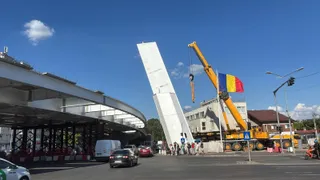 O macararie galbenă ridică o structură albă verticală pe un drum urban lângă un pasaj rutier și câteva mașini circulând.
