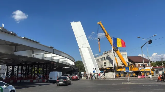 O macararie galbenă ridică o structură albă verticală pe un drum urban lângă un pasaj rutier și câteva mașini circulând.