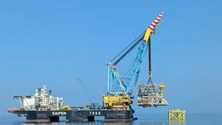 O platformă de lucru cu o macara albastră și galbenă ridică o structură metalică deasupra apei sub un cer senin.
