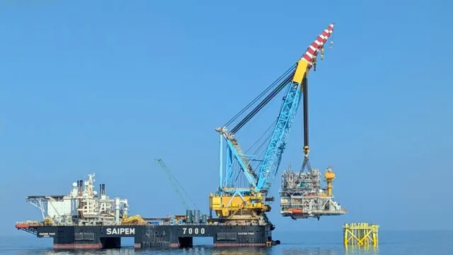 O platformă de lucru cu o macara albastră și galbenă ridică o structură metalică deasupra apei sub un cer senin.