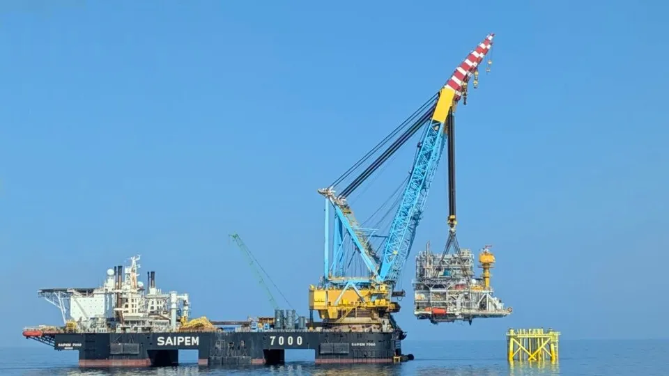 O platformă de lucru cu o macara albastră și galbenă ridică o structură metalică deasupra apei sub un cer senin.
