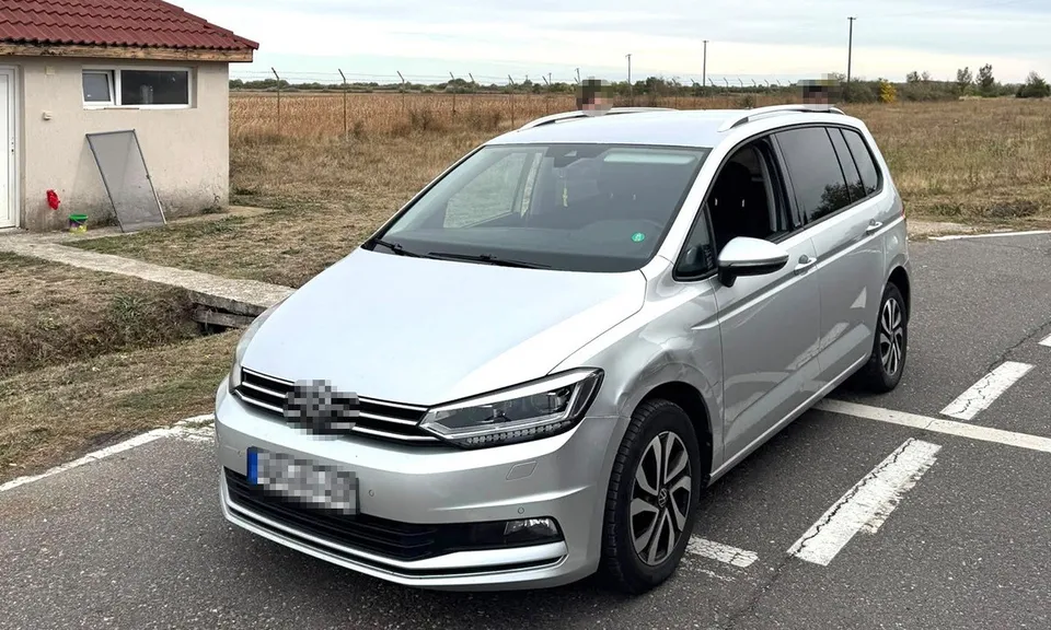 Un autoturism argintiu este parcat pe un drum asfaltat lângă un câmp uscat și o clădire mică cu acoperiș de țiglă roșie.