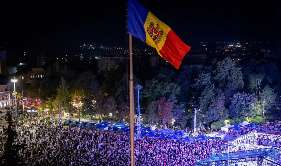 O mulțime de oameni este adunată într-o zonă deschisă noaptea, sub un steag tricolor care flutură pe un stâlp înalt.