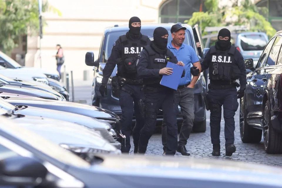 Trei persoane purtând măști negre și vesta cu inscripția B.S.I.J. se află lângă un vehicul alb și un bărbat în tricou albastru.