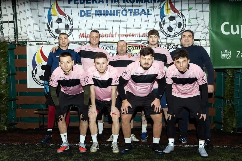Echipa de minifotbal pozată în fața unei poțe, purtând tricouri roz cu o bandă neagră orizontală și pantaloni scurți negri.
