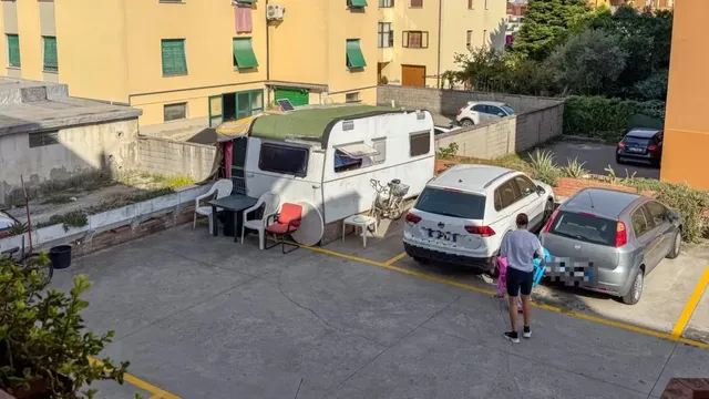 O rulotă albă cu acoperiș verde este parcată într-o curte lângă un vehicul alb, un vehicul gri și un bărbat care merge pe asfalt.
