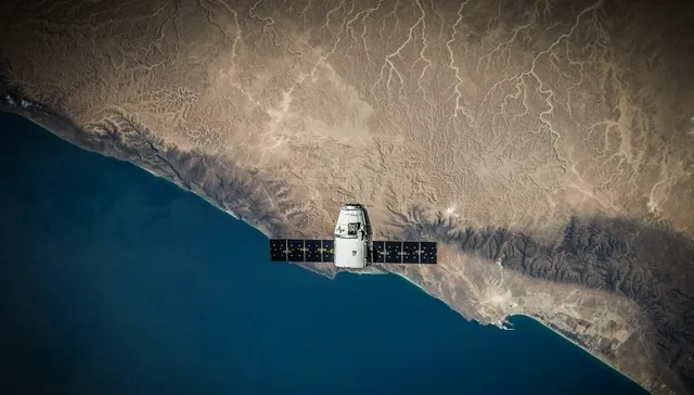 Un satelit cu panouri solare negre planează deasupra unei linii de coastă dintre un ocean albastru și un teren muntos de culoare maro.