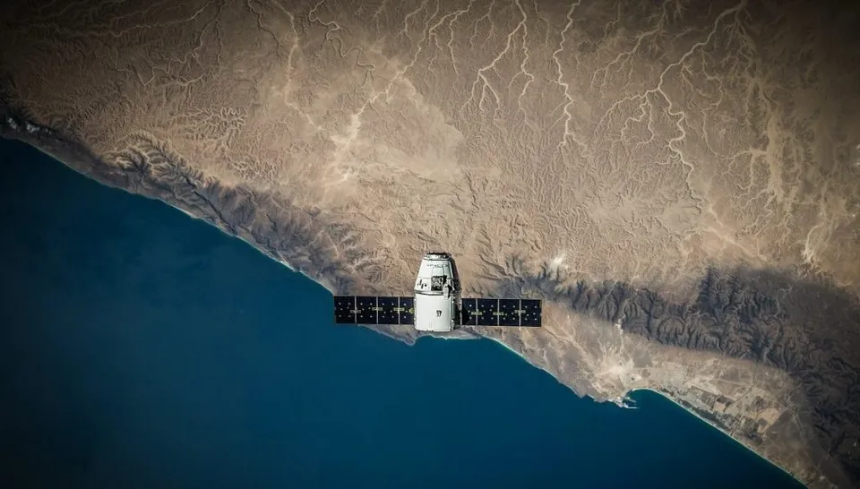 Un satelit cu panouri solare negre planează deasupra unei linii de coastă dintre un ocean albastru și un teren muntos de culoare maro.