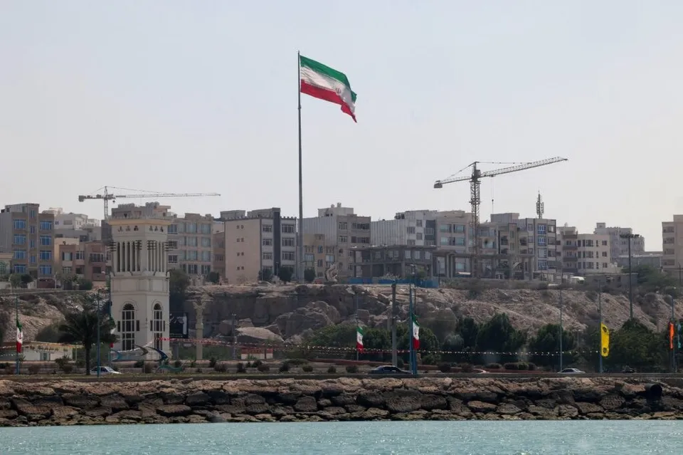 Un steag iranian flutură pe un stâlp înalt deasupra unor clădiri urbane și a unui mal stâncos lângă apă.