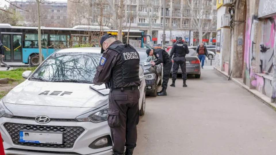 Unii agenți în uniformă neagră cu inscripția Politia Locala verifică un vehicul alb parcat pe marginea unui drum urban lângă un autobuz albastru.