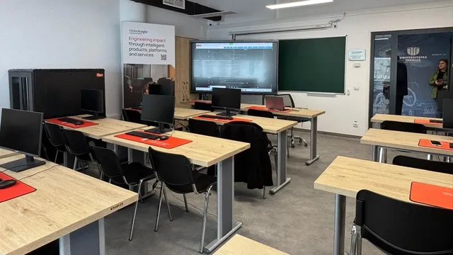 O sală de clasă cu mai multe birouri de lemn deschise, monitoare negre, scaune negre și un ecran mare montat pe perete.