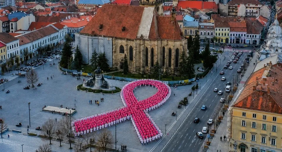 O mulțime de persoane purtând haine roz formează o panglică mare pe o piață pavată lângă o biserică cu acoperiș roșu.