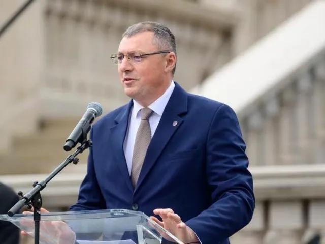 Un bărbat cu ochelari și costum albastru stă în spatele unui podium transparent și vorbește la un microfon.