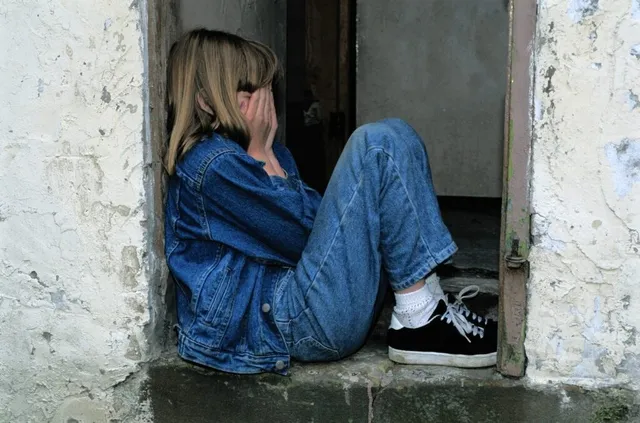 O persoană cu părul blond, purtând o geacă de denim și pantaloni de denim, stă ghemuită într-o deschidere de perete cu fața acoperită de mâini.