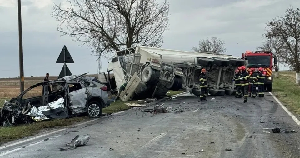 Un autoturism gri distrus stă pe marginea drumului lângă un autotren alb răsturnat, în timp ce mai mulți pompieri stau lângă vehicule.