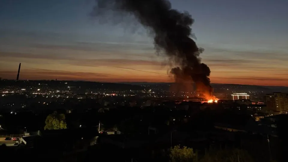 Un nor gros de fum negru se ridică deasupra unui oraș în timpul amurgului, deasupra unor clădiri și luminilor urbane.