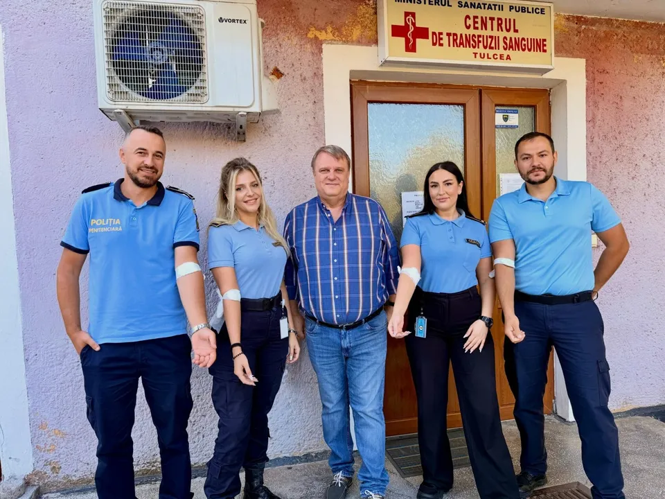 Cinci persoane stau în rând în fața unei uși, patru purtând tricouri albastre și pantaloni închizi, iar unul purtând o cămașă în dungi și blugi.