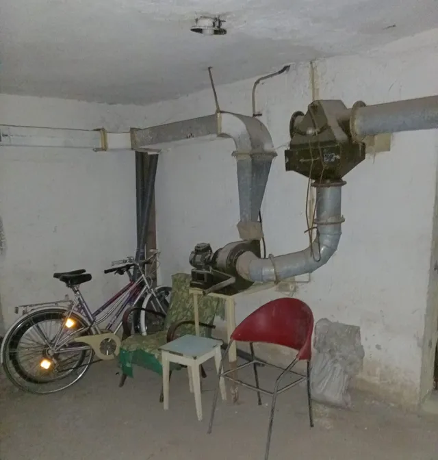 Într-o încăpere cu pereți albi, se află o bicicletă, un scaun roșu, un scaun verde, o masă mică și conducte metalice suspendate.