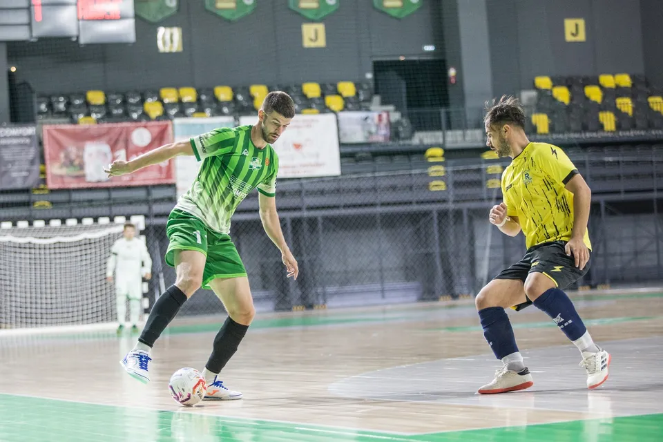 Un jucător în echipament verde și alb conduce mingea pe un teren de futsal, fiind urmărit de un adversar în echipament galben și negru.
