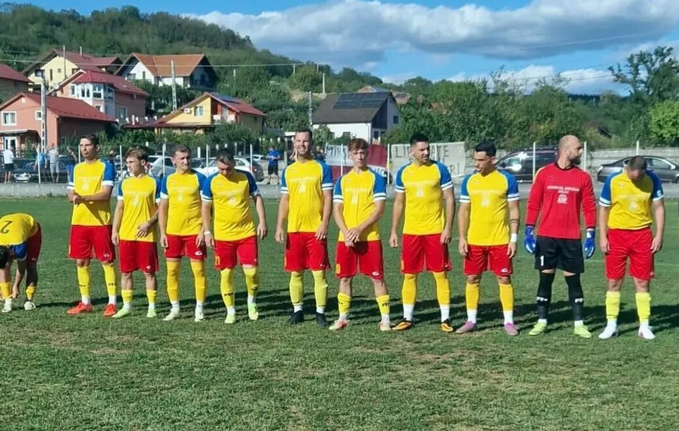 O echipă de fotbaliști îmbrăcați în echipamente galben și albastru stau în linie pe un teren de iarbă verde sub un cer înnorat.