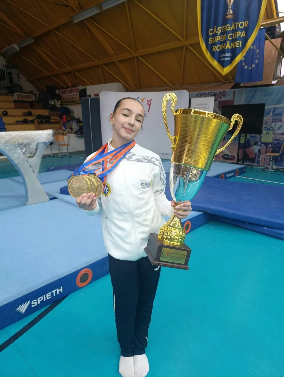 O fată într-un costum alb și pantaloni închiși ține o medalie aurie și un trofeu mare de culoare aurie într-o sală de sport.