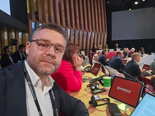 Un bărbat cu ochelari și sacou negru face un selfie într-o sală de conferințe cu oameni așezați la mese cu laptopuri și tablete.