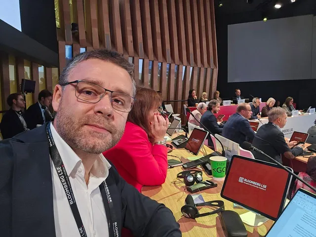 Un bărbat cu ochelari și sacou negru face un selfie într-o sală de conferințe cu oameni așezați la mese cu laptopuri și tablete.