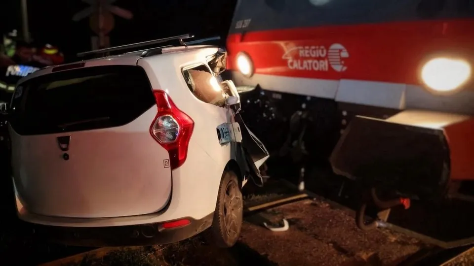 Un autoturism alb avariat se află pe șine lângă locomotiva Roșie, cu iluminarea aprinsă.