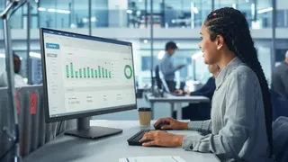O femeie cu părul împletit stă la un birou și privește un monitor care afișează grafice și date într-un birou modern.