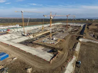 O vedere aeriană prezintă un șantier cu excavări mari, macarale de construcție și drumuri de pământ sub un cer senin.