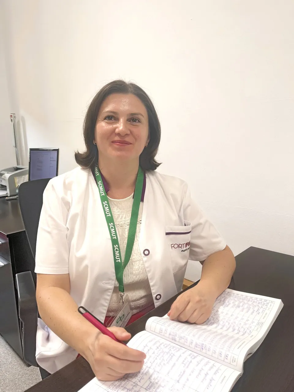 O femeie cu halat alb și lanyard verde stă la birou ținând un stilou roșu și un registru deschis.