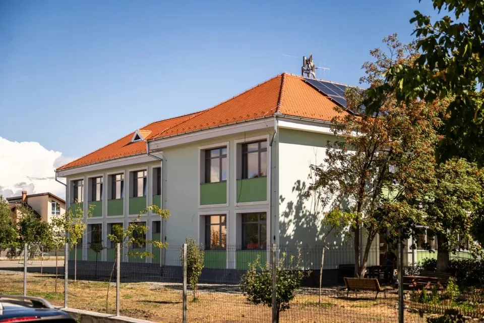 O clădire cu fațadă verde și acoperiș portocaliu, dotată cu panouri solare pe acoperiș, este înconjurată de un gard și vegetație sub un cer senin.
