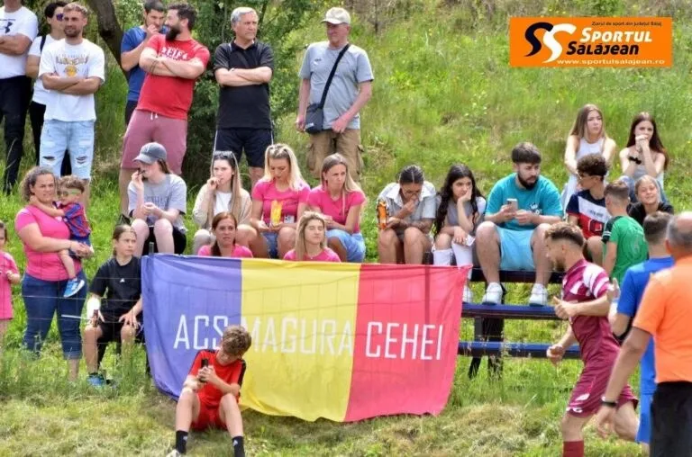 Un grup de persoane stă pe o bancă și pe iarbă lângă un steag colorat cu textul ACS Magura Cehei.