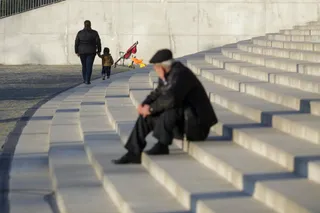 Un bărbat cu pălărie neagră stă așezat pe niște trepte de beton, în timp ce în fundal un adult și un copil merg pe un drum curbat.