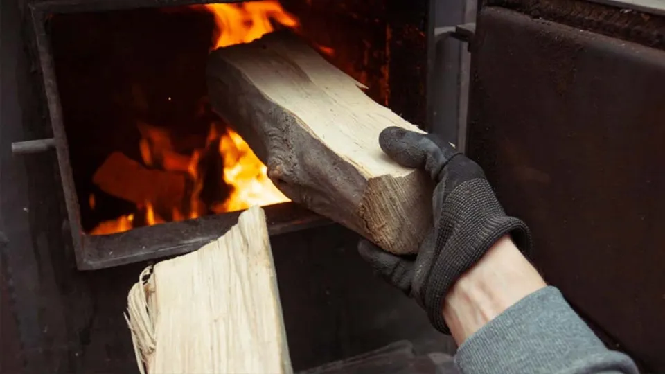 O mână cu mănușă neagră ține o bucată de lemne tăiată și o introduce într-o cuptor cu flăcări aprinse în interior.