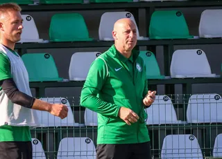 Un bărbat cu o vestă verde și un alt bărbat cu o cămașă albă stau în fața unor scaune de stadion verzi și albe.