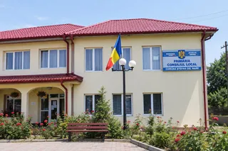 O clădire cu acoperiș roșu, un stâlp cu două globuri albe și un banc de grădină situat în fața unei vegetații cu flori roșii.