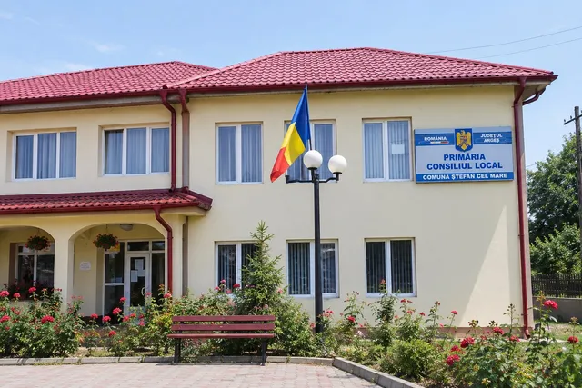 O clădire cu acoperiș roșu, un stâlp cu două globuri albe și un banc de grădină situat în fața unei vegetații cu flori roșii.