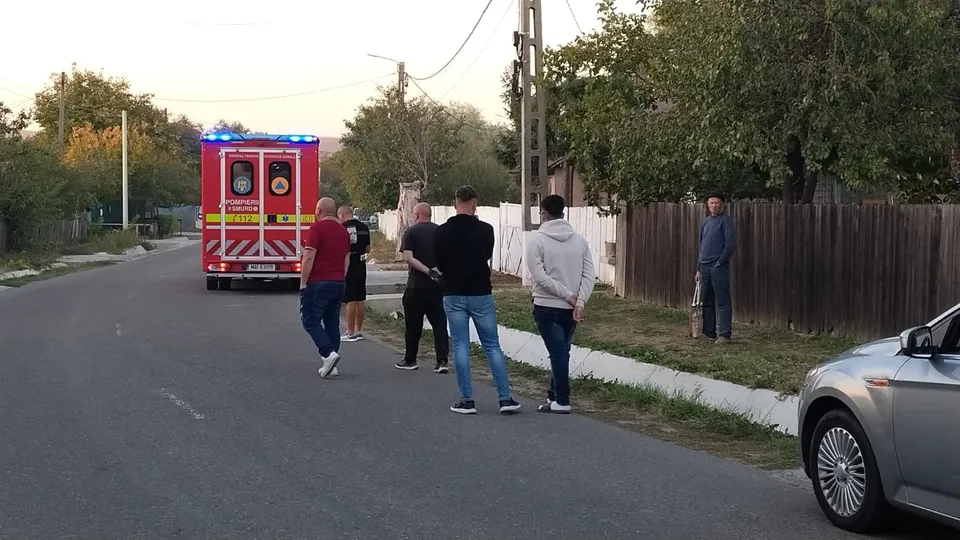 O ambulanță cu luminile aprinse stă pe marginea unui drum, în timp ce un grup de oameni stă pe asfalt lângă ea.